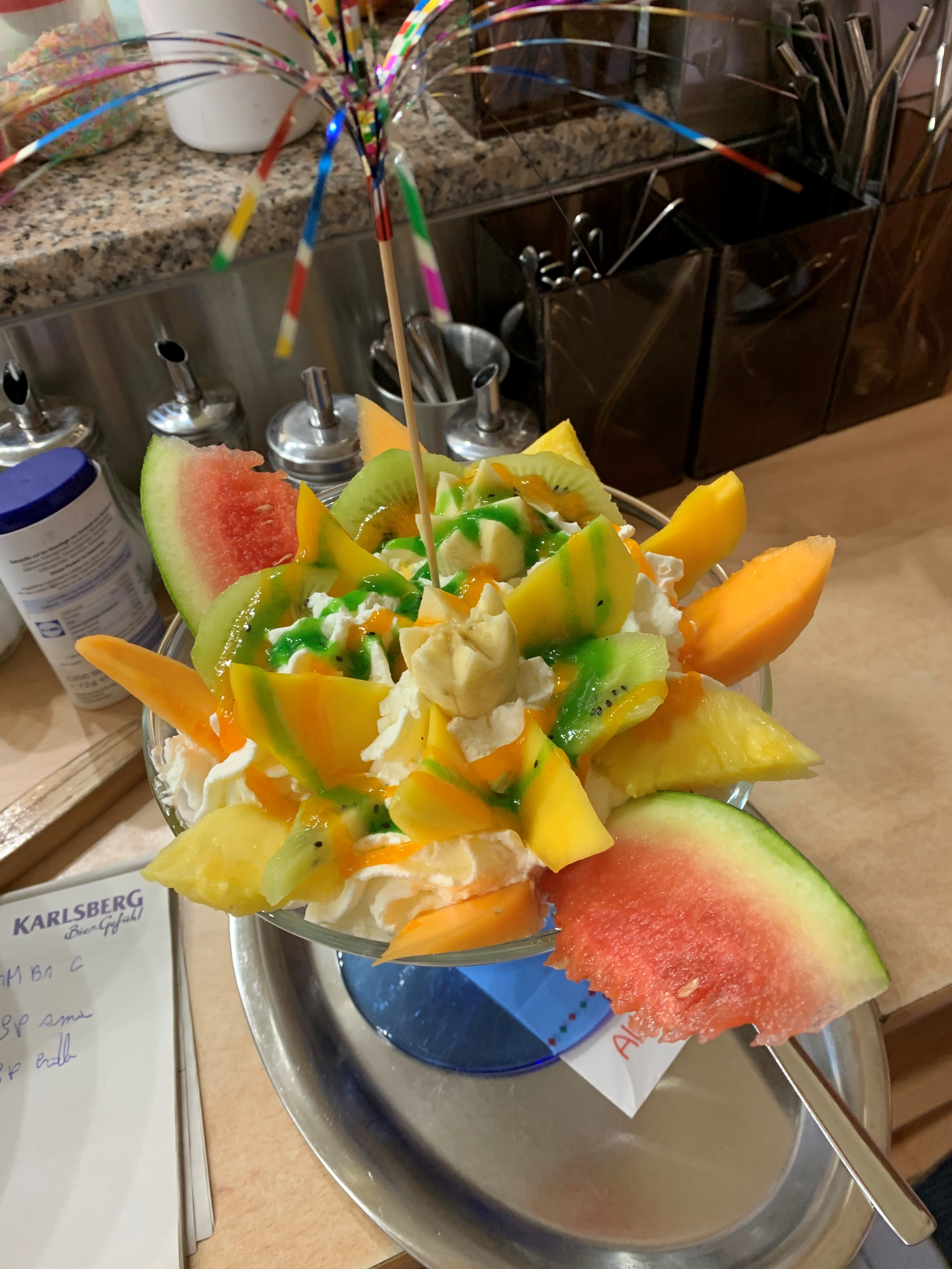 Bunt dekorierter Eisbecher mit verschiedenen frischen Früchten wie Wassermelone, Kiwi und Mango, garniert mit Schlagsahne und grünem Sirup, präsentiert auf einer Theke.
