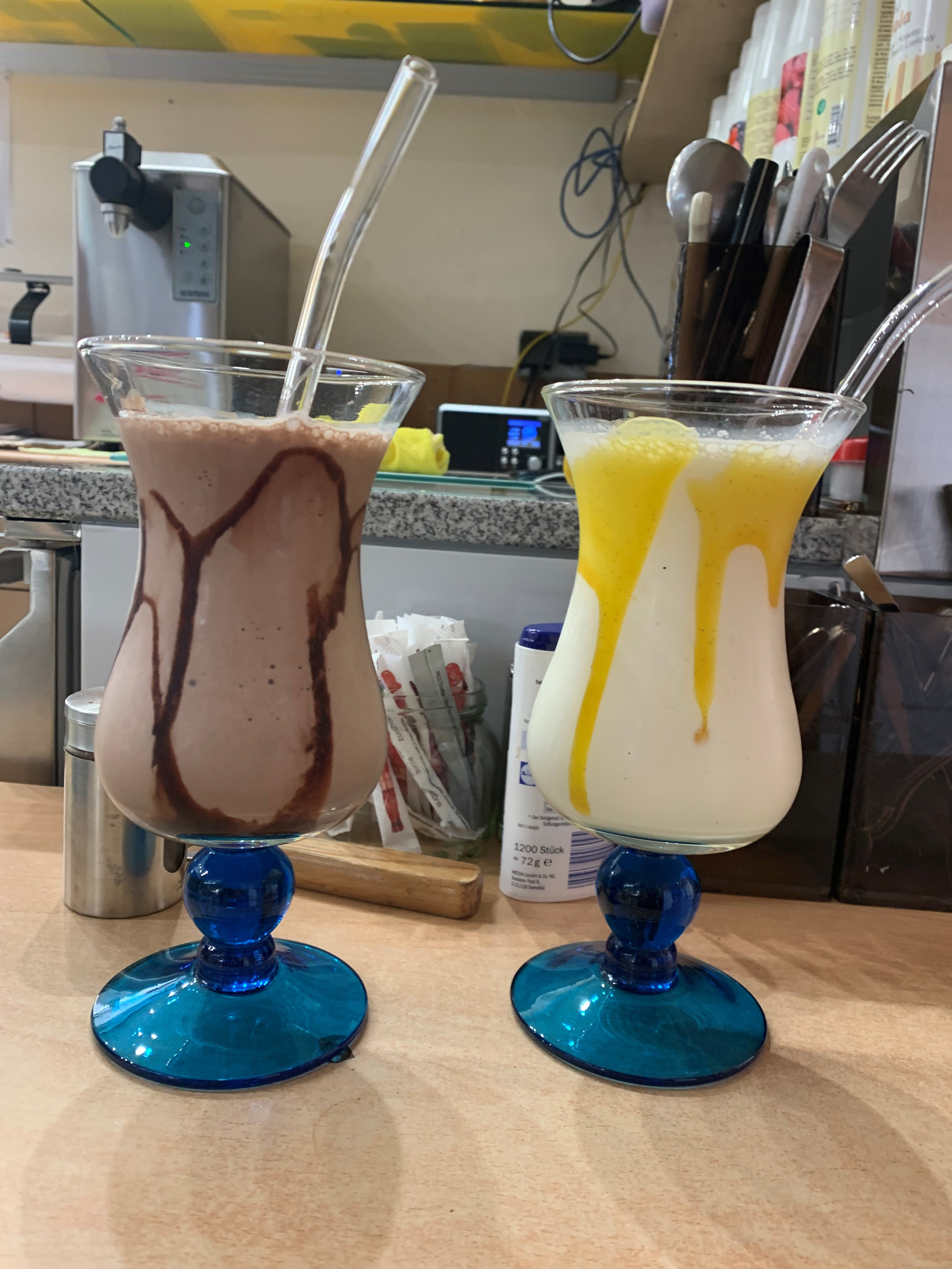 Zwei Milchshakes mit Glasstrohhalmen, links ein Schokoladenmilchshake mit Schokoladensauce, rechts ein Vanillemilchshake mit Mangosauce, beide in Gläsern mit blauem Fuß, auf einer Café-Theke.