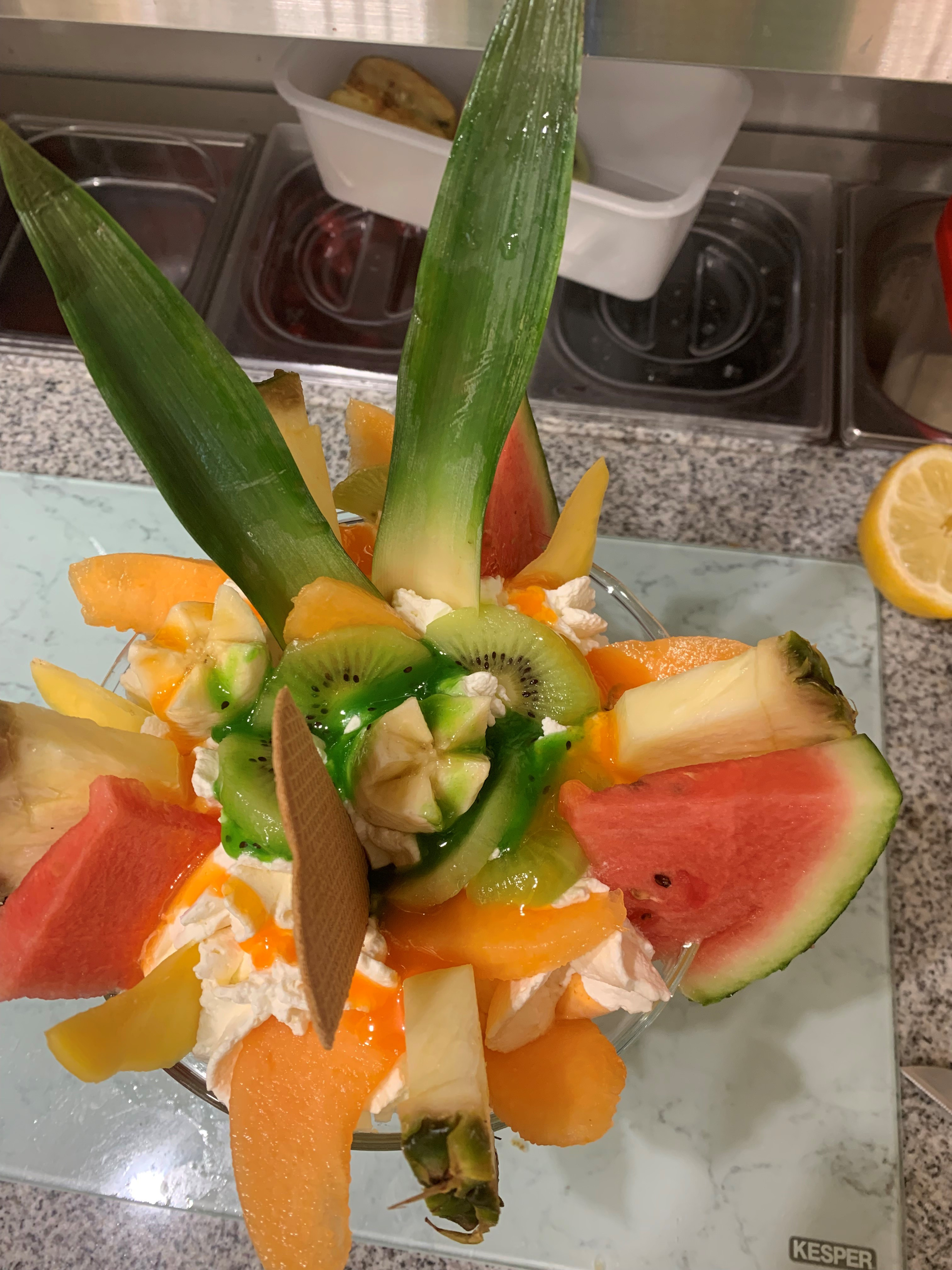 Exotischer Fruchtbecher mit frischen Kiwis, Wassermelonenstücken, Mango und Ananas, dekoriert mit grüner Kiwisauce und zwei Waffelröllchen, serviert auf einer hellen Marmorplatte in der Eisdiele.